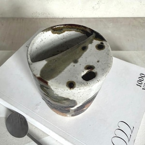 Könnte beinhalten: Eine handgefertigte Keramikvase mit einem einzigartigen Design. Die zylindrische Vase hat einen weißen Sockel, verziert mit braunen, grünen und schwarzen organischen Mustern. Sie hat eine gebogene Öffnung und ein kleines Loch. Die Vase steht auf einem Buch.