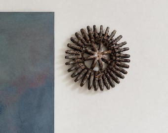 May include: A circular wall sculpture featuring a dark brown, textured, gear-like form with numerous radiating spokes. The object has a weathered, metallic appearance with subtle highlights of gold and bronze tones along its edges and center.