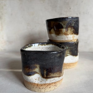 May include: A stack of three handmade ceramic cups. The cups have a dark brown glaze at the top, transitioning to a white band, and ending with a speckled tan base. The cups are likely used for drinking.
