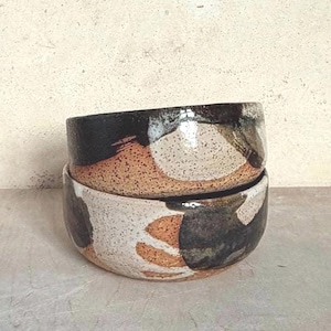 Może przedstawiać: Dwie ceramiczne miski ułożone w stos z nakrapianą, ziemistą estetyką. Miski mają mieszankę kolorów, w tym czarny, biały i brązowy, z błyszczącym wykończeniem. Projekt jest abstrakcyjny i organiczny, odpowiedni do serwowania żywności lub jako elementy dekoracyjne.