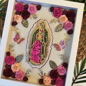 Könnte beinhalten: Ein weiß gerahmtes Bild mit einer Papierschnittarbeit der Jungfrau Maria in einem goldenen und rosafarbenen Rahmen, umgeben von rosa, burgunderroten und pfirsichfarbenen Papierblumen. Die Blumen sind um das Bild herum und in den Ecken des Rahmens angeordnet.