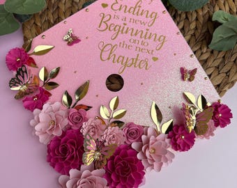 May include: Pink graduation cap decorated with paper flowers, gold butterflies, and leaves. The cap has the text "Every Ending is a new Beginning onto the next Chapter" in gold script. The flowers are shades of pink and fuchsia.