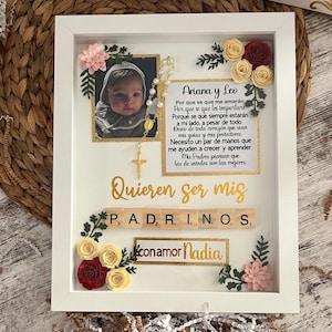 May include: A white picture frame with a gold border and a photo of a baby inside. The frame is decorated with paper flowers in shades of red, yellow, and pink. The text "Ariana y Leo" is written in black lettering on a white background. The text "Quieren ser mis P, A, D, R, I, N, O, S, conamor Nadia" is written in gold lettering on a white background.