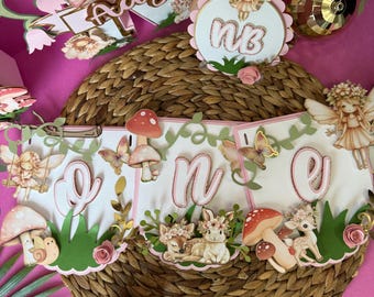 May include: A whimsical, pink and white banner with the word "one" and fairy-themed decorations. The banner includes mushroom and butterfly accents, along with a woven basket and other decorative elements. The overall theme is a birthday celebration.