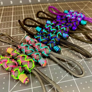 May include: Close-up of several handmade paracord keychains in various colors. The keychains feature intricate knotwork in black, gray, purple, teal, pink, and neon green. Each keychain has a loop for easy attachment.