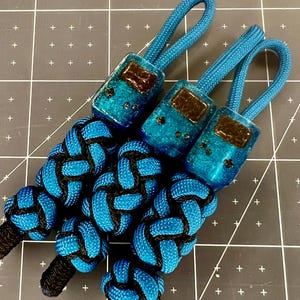 May include: Three handmade zipper pulls with blue and black braided knots. Each pull features a blue resin charm with a dark brown design and a matching blue cord loop. The background is a gray cutting mat with white grid lines.