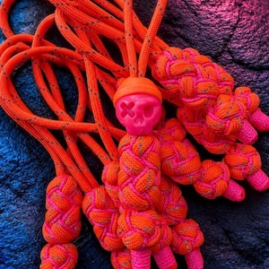 May include: A set of bright orange and pink paracord zipper pulls featuring a central skull bead with an orange beanie. The cords are woven into intricate knots with reflective silver flecks throughout the material, creating a textured look.
