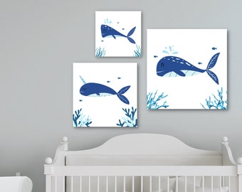 May include: A set of three blue and white framed prints featuring illustrations of whales and a narwhal swimming in the ocean. The prints are displayed on a white wall above a white crib.
