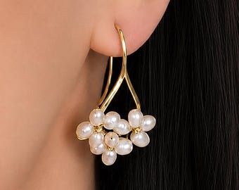 May include: A gold-toned earring featuring a curved wire hook that supports a cluster of small, white, irregular-shaped freshwater pearls arranged in a floral-like formation. The metal hook has a smooth, polished finish and a V-shaped design.