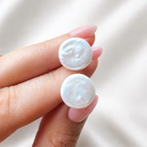 May include: Two round, flat, white coin-shaped pearls with a lustrous, iridescent surface. Each pearl features a smooth, circular silhouette with subtle natural texture and a high-gloss finish, reflecting light across their polished, pearlescent surfaces.