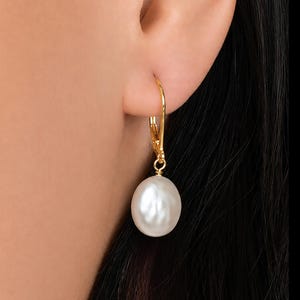 May include: A gold leverback earring featuring a single, smooth, white, oval-shaped pearl drop. The polished gold metal hook secures the pearl, which hangs vertically from the earlobe, showcasing a lustrous, reflective surface and a classic design.