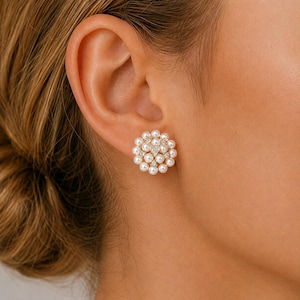 May include: A circular stud earring featuring a cluster of small, round, white pearls arranged in a floral pattern. The center of the earring contains a small, clear, sparkling stone, and the pearls are set against a gold-toned metal base.