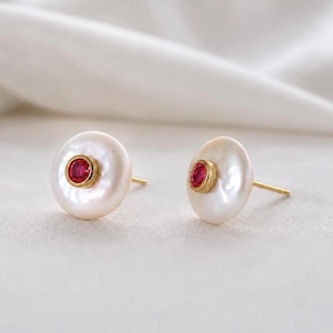 May include: A pair of stud earrings featuring round, white, iridescent pearls. Each pearl is centered with a small, circular, vibrant red gemstone set in a polished gold-tone metal bezel. The earrings include thin gold-tone posts for attachment.