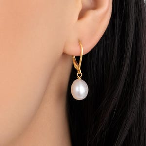 May include: A gold leverback earring featuring a single, smooth, round white pearl drop. The earring is suspended from a gold wire mechanism that secures through the earlobe, showcasing a polished metallic finish and a lustrous pearl surface.