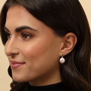 Puede incluir: Una mujer con una camisa negra y un pendiente de perla con un acento de piedra verde.