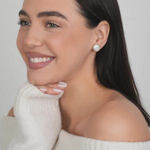 May include: A round stud earring featuring a central iridescent white pearl-like stone set within a thin, polished gold-tone metal rim. The earring is circular in shape and sits flush against the earlobe, showcasing a smooth, reflective surface.