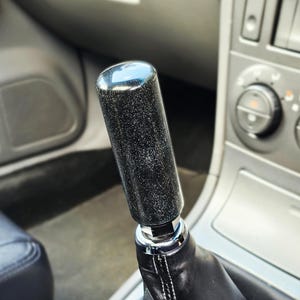 May include: A cylindrical gear shift knob with a black finish and fine silver glitter particles, mounted on a black leather shift boot with visible white stitching, positioned within the centre console of a vehicle interior.