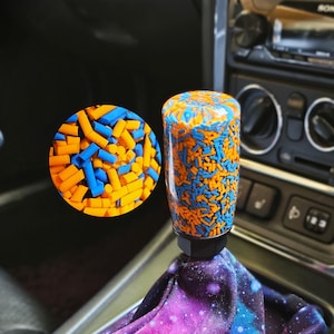May include: A cylindrical gear shift knob filled with small orange and blue plastic sprinkles, attached to a black base and a fabric shift boot featuring a purple, pink, and blue galaxy print with white star-like speckles throughout the fabric.