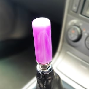 Peut inclure: Un pommeau de levier de vitesse en forme de spirale violet et blanc avec un soufflet en cuir noir. Le pommeau est installé dans une voiture.
