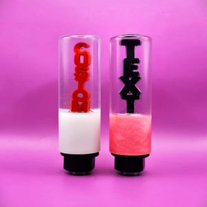 May include: Two cylindrical gear shift knobs with clear glass bodies and black bases. The left knob contains a white liquid base and red vertical text reading CUSTOM. The right knob contains a pink liquid base and black vertical text reading TEXT.
