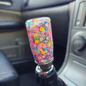 Può includere: Un pomello del cambio colorato con un design floreale. Il pomello cilindrico è ricoperto da un motivo di piccoli fiori dai colori vivaci in rosa, giallo, blu e verde. È attaccato a un soffietto del cambio in pelle nera.