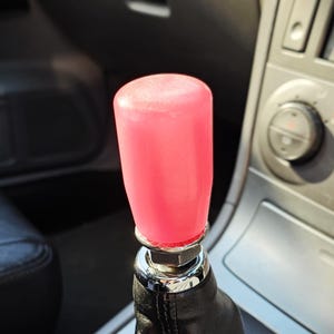 May include: A pink gear stick knob with a chrome base, set within a car's interior. The knob is cylindrical and appears translucent. The gear stick boot is made of black leather.
