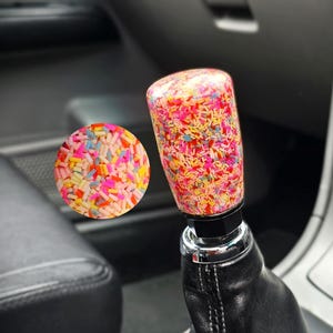 Op de afbeelding: Een aangepaste auto versnellingspookknop met een kleurrijk hagelslagontwerp. De cilindrische knop is gevuld met een mix van roze, gele, blauwe en oranje hagelslag, tegen een heldere achtergrond.