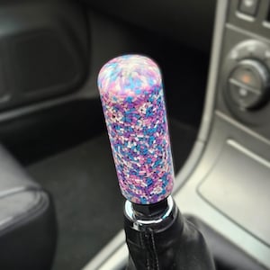 Op de afbeelding: Een kleurrijke, cilindrische versnellingspookknop met een mozaïekpatroon van kleine, rechthoekige stukjes in roze, paars en blauw. De knop is gemonteerd op een zwarte leren versnellingspookhoes, tegen een vage auto-interieur.