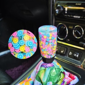 May include: A cylindrical gear shift knob filled with small, colourful, translucent citrus fruit slices in shades of pink, yellow, and blue, attached to a fabric shift boot featuring a vibrant tropical floral pattern with green leaves and pink flowers.