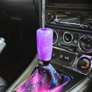 May include: A cylindrical gear shift knob with a translucent purple finish containing fine glitter, attached to a fabric shift boot featuring a vibrant galaxy print with shades of pink, purple, and blue, installed in a vehicle center console.