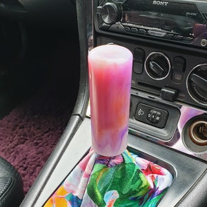 May include: A pink and white swirl resin gear shift knob with a floral patterned gear shift boot. The boot has a tropical print with pink, green, yellow, and blue flowers.