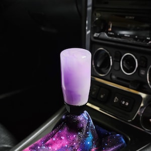 May include: A cylindrical gear shift knob with a smooth, marbled lavender and white finish, attached to a black base. The gear shift boot features a vibrant galaxy print with shades of pink, purple, and blue, set within a car interior console.