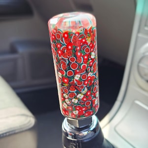 May include: A custom car gear stick knob. The clear, cylindrical knob is filled with red, white, and green fruit slices. It is mounted on a black leather shift boot, with a chrome base.