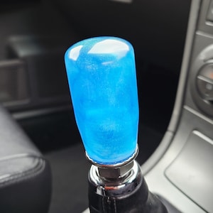 May include: A custom blue shift knob with a glittery, translucent appearance. The knob is cylindrical with a rounded top and is mounted on a black leather shift boot. The surrounding interior is grey and black.