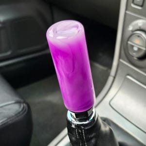 May include: A custom car gear stick knob featuring a swirling purple and white pattern. The cylindrical knob is attached to a black leather gear stick gaiter, set within a car's interior. The knob has a glossy, reflective surface.