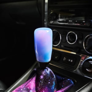 May include: A cylindrical gear shift knob with a smooth, translucent blue and pink marbled finish, attached to a black base. Below the knob is a fabric gear shift boot featuring a vibrant galaxy print with purple, pink, and blue nebula patterns.