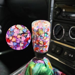 May include: A cylindrical gear shift knob filled with a colorful assortment of small heart-shaped confetti pieces in shades of pink, blue, yellow, and purple. The knob is attached to a fabric shift boot featuring a vibrant floral print with hibiscus flowers.