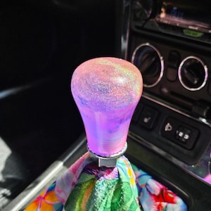 May include: A custom car gear stick knob with a swirling pink and purple design, embedded with glitter. The knob sits atop a floral patterned gear stick gaiter with a colourful mix of flowers and leaves.