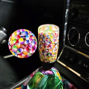 Può includere: Un pomello del cambio cilindrico con un corpo in resina trasparente riempito da un fitto motivo di piccole forme multicolori simili a petali, fissato a una base nera e a una cuffia in tessuto con una vivace stampa floreale tropicale.