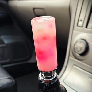 Op de afbeelding: Een aangepaste auto versnellingspookknop met een roze en wit doorschijnend ontwerp. De cilindrische knop is gemonteerd op een zwarte leren schakelhoezen, met een chromen basis. Het interieur van de auto is zichtbaar op de achtergrond.
