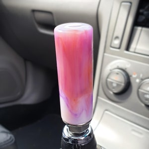 May include: A pink and white swirl resin gear shift knob with a chrome base. The knob is attached to a black leather gear shift boot.