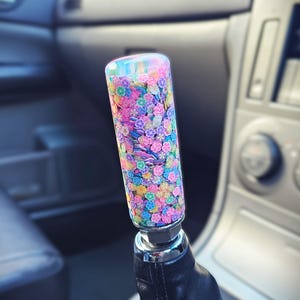 Könnte beinhalten: Ein farbenfroher Schaltknauf mit floralem Design. Der Knopf ist zylindrisch und mit kleinen, mehrfarbigen Blumen bedeckt. Der Hintergrund zeigt das Innere eines Autos.