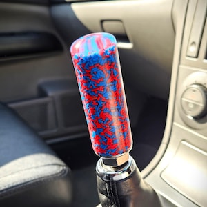 May include: A custom shift knob with a colourful design. The cylindrical knob has a clear resin exterior filled with red and blue sprinkles. It is mounted on a chrome base and a black leather shift boot, inside a car interior.