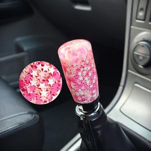 May include: A cylindrical gear shift knob made of clear resin filled with numerous small pink and white star-shaped confetti pieces. The knob is attached to a black leather gear shift boot, with a circular inset showing a close-up of the stars.