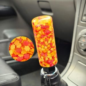 Pode incluir: Um botão de mudança de marcha de carro personalizado com um design vibrante e colorido. O botão é cilíndrico e preenchido com pequenos confetes em forma de flor em tons de vermelho, laranja e amarelo. Está preso a uma bota de mudança de marcha de couro preto.