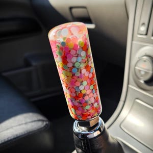 May include: A vibrant shift knob with a clear, cylindrical design filled with confetti in various colours. The knob is attached to a black leather shift boot in a car interior.