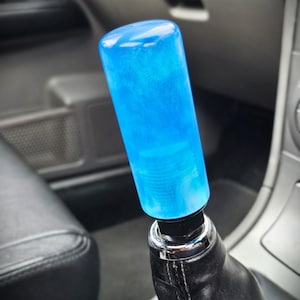 Op de afbeelding: Een helderblauwe versnellingspookknop. De cilindrische knop is gemaakt van een doorschijnend materiaal en is gemonteerd op een zwarte leren versnellingspookhoes. De binnenkant van een auto is zichtbaar op de achtergrond.