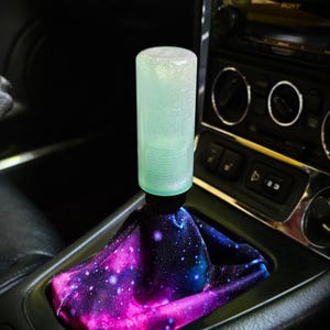 Könnte beinhalten: Ein zylindrischer Schaltknauf mit einer durchscheinenden, hellgrünen, glitzernden Oberfläche auf einem schwarzen Sockel. Die Schalthebelmanschette besteht aus Stoff mit einem lebendigen Galaxie-Druck in tiefem Violett, Blau und leuchtendem Pink.
