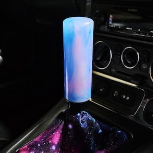 May include: A cylindrical gear shift knob with a smooth, translucent blue and pink marbled finish, attached to a black base. The knob is paired with a fabric shift boot featuring a vibrant galaxy print with purple, pink, and blue nebula patterns.
