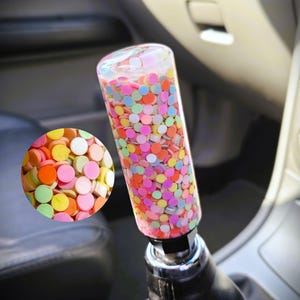 May include: A car gear shift knob with a clear, cylindrical design filled with colorful, circular confetti. The confetti includes shades of pink, orange, yellow, green, and blue. The knob is mounted on a black leather shift boot.